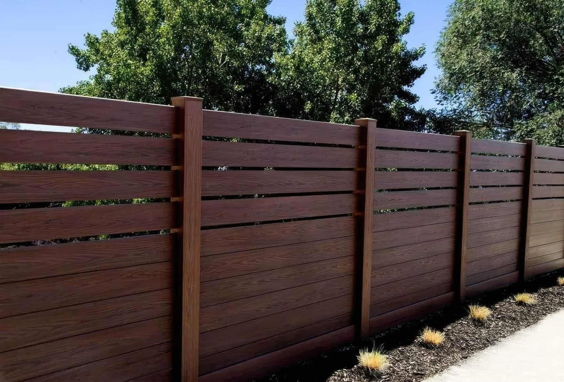Brown vinyl fence with semi-privacy horizontal slats in front of large trees and behind concrete walkway and small plants