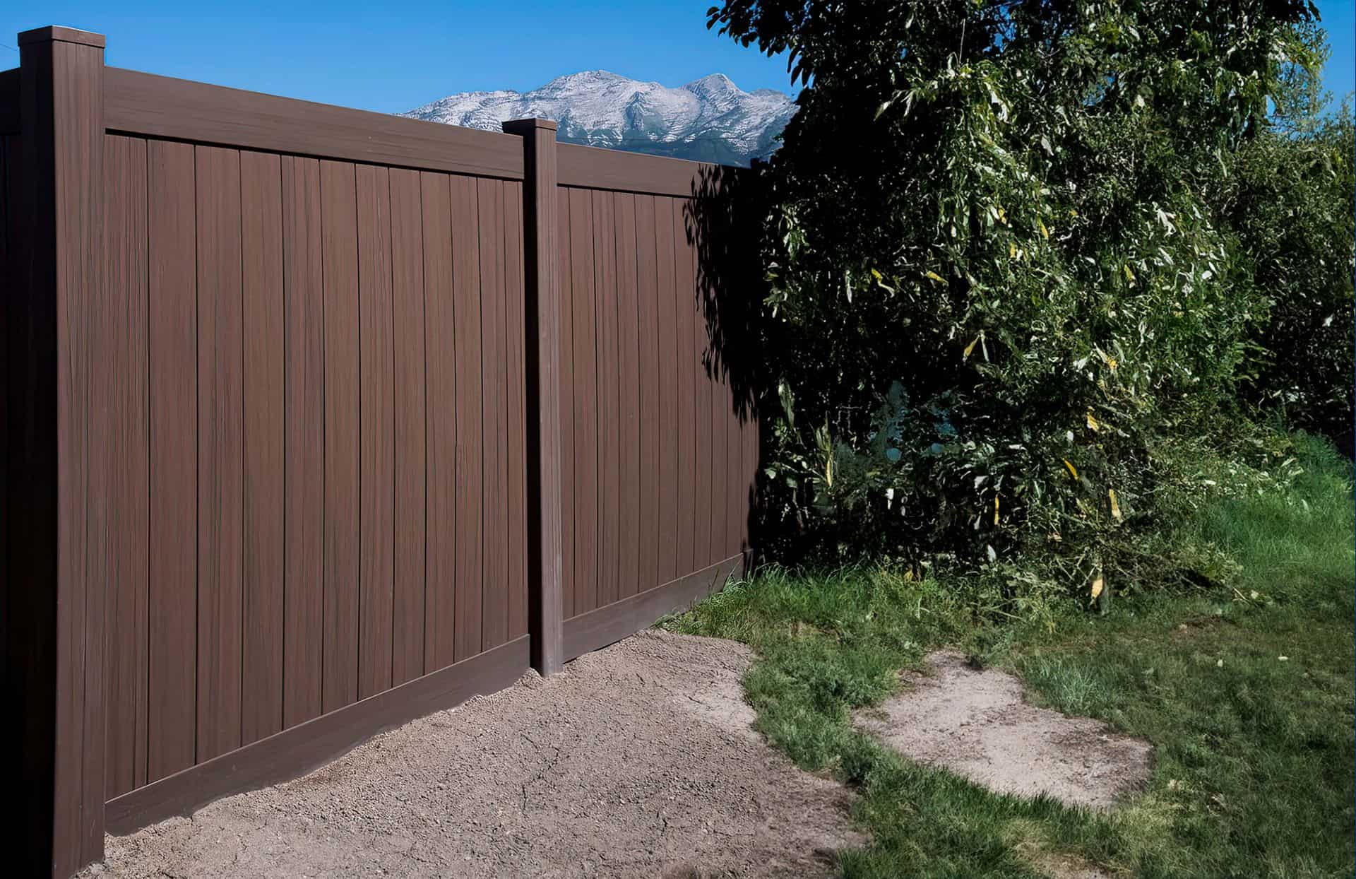 Dark sequoia vinyl fence with vertical slats behind large tree and grassy lawn, with dirt walkway.