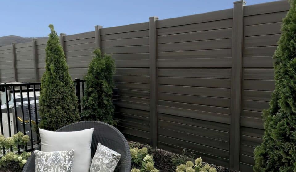 Dark sequoia vinyl fence with horizontal slats next to small trees and metal fence, along with small plants and bushes in the garden.