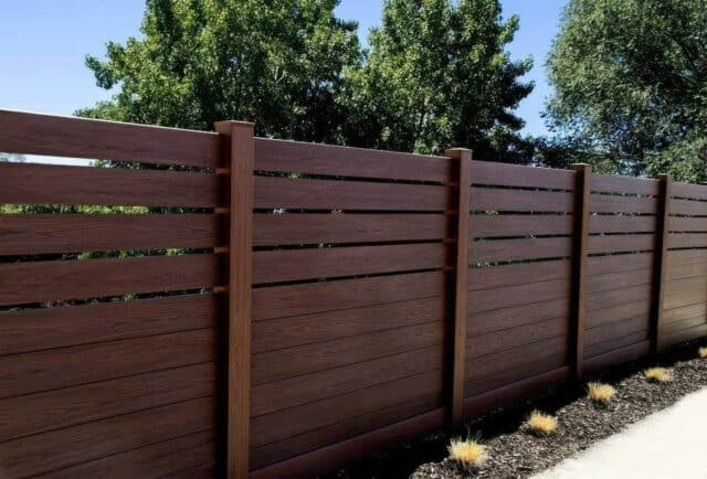 Dark sequoia vinyl fence beside concrete sidewalk with large trees in the background and a clear blue sky.