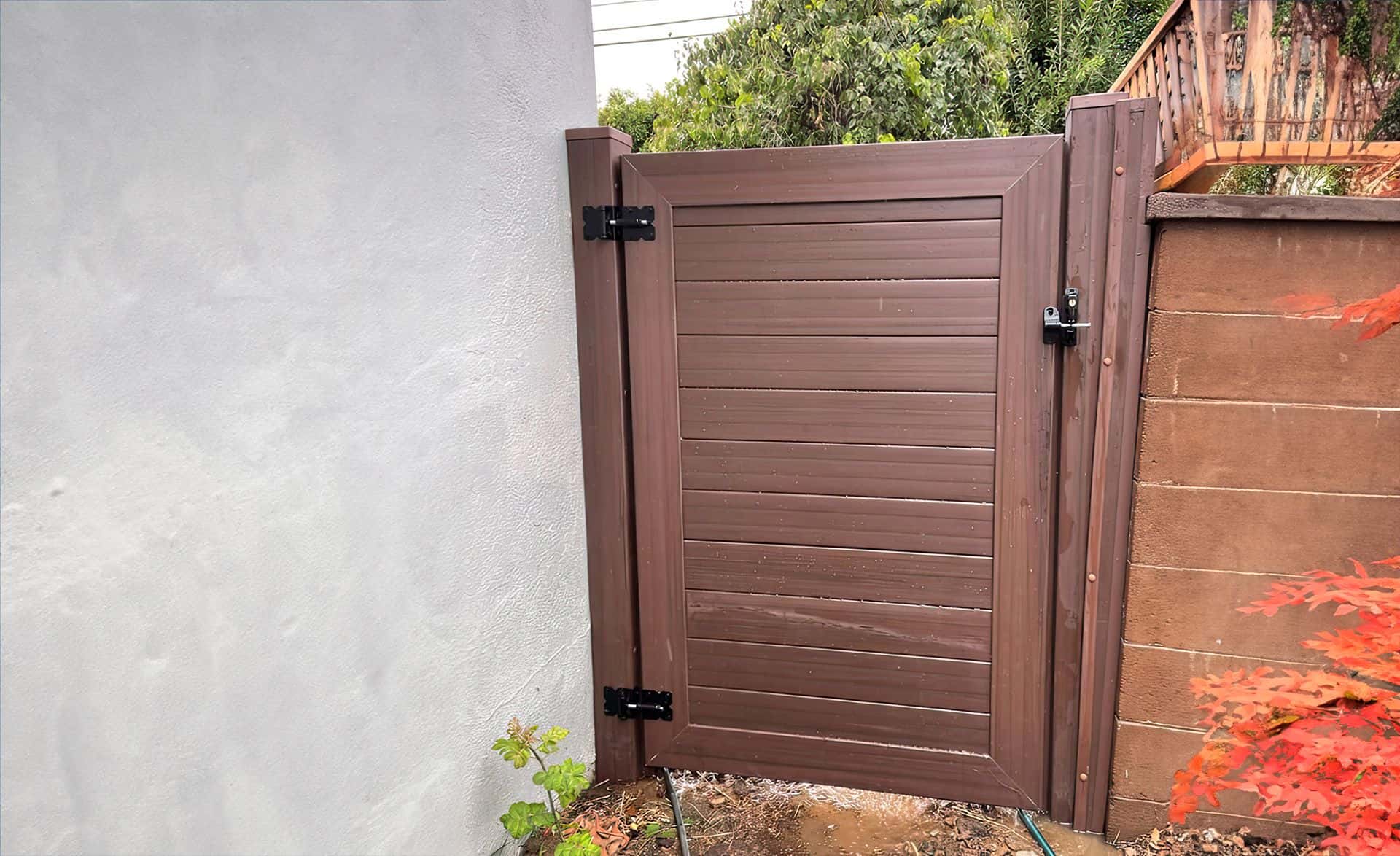 Dark sequoia vinyl fence gate with black hinges next to red brick boundary wall, leading into backyard with garden and trees.