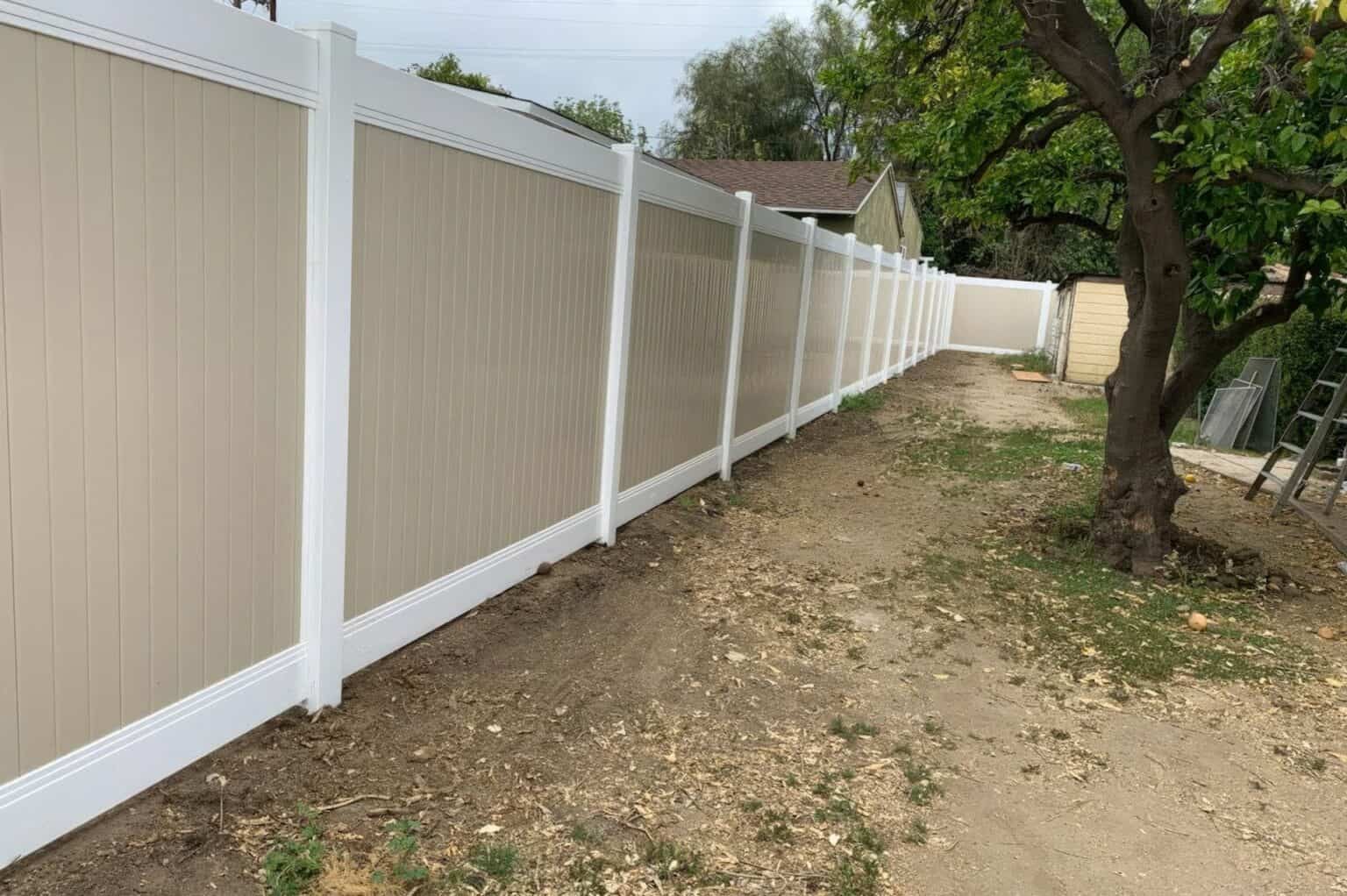 Tan vinyl fence creating boundary around modern suburban home with large tree in the backyard and other houses in the back.