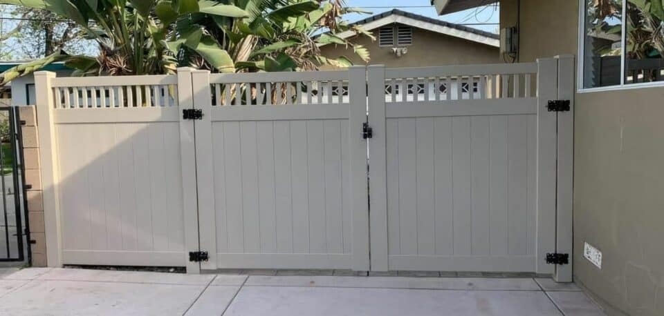 Tan vinyl fence with gates to driveway behind small trees and right beside neighbor's metal gate