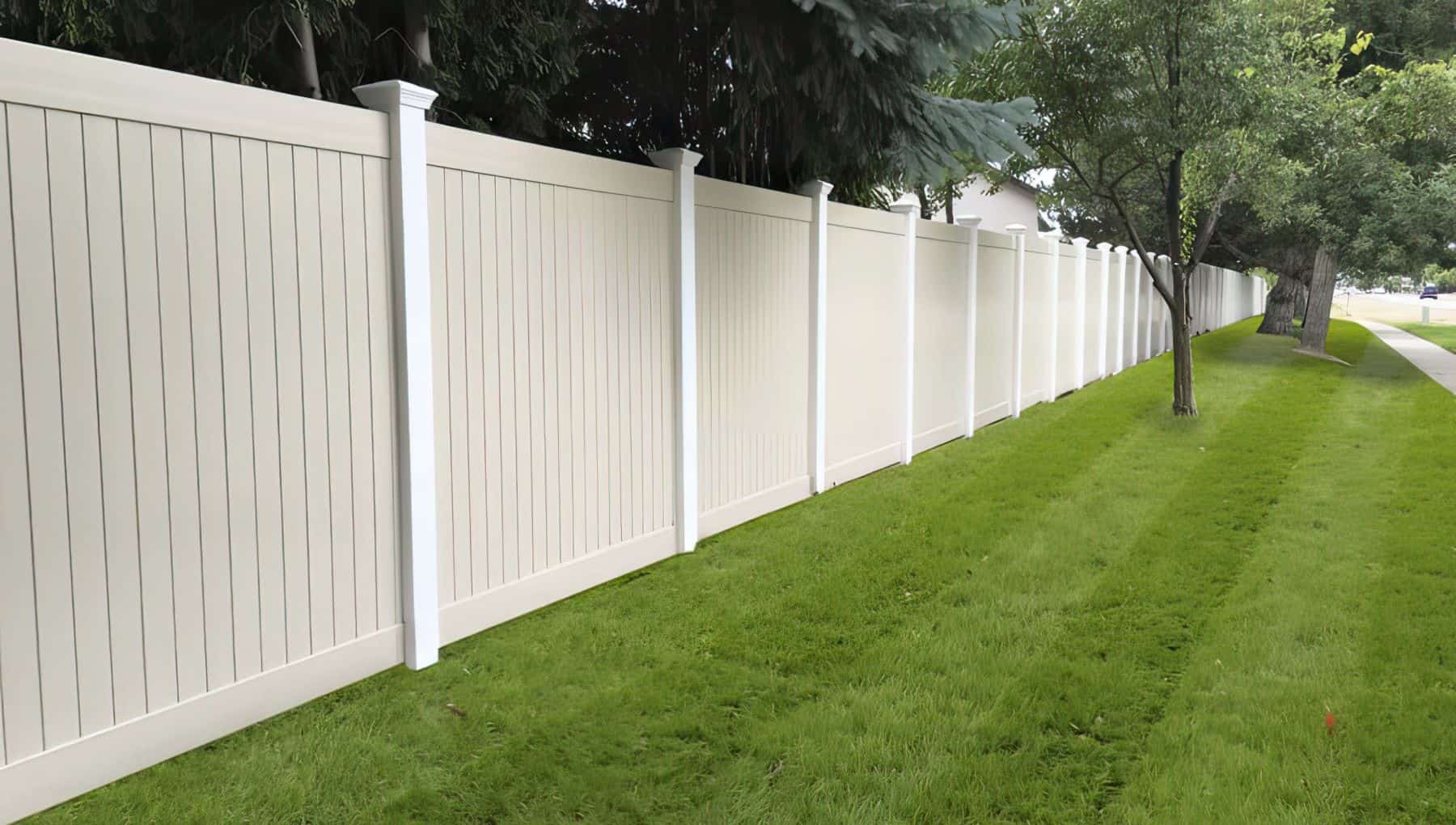 Tan vinyl fence in front of small trees on a manicured grassy lawn with bigger trees in the background.