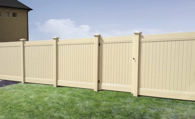 Tan vinyl fence with gate on grassy lawn with a clear blue sky in the background next to a small townhouse.
