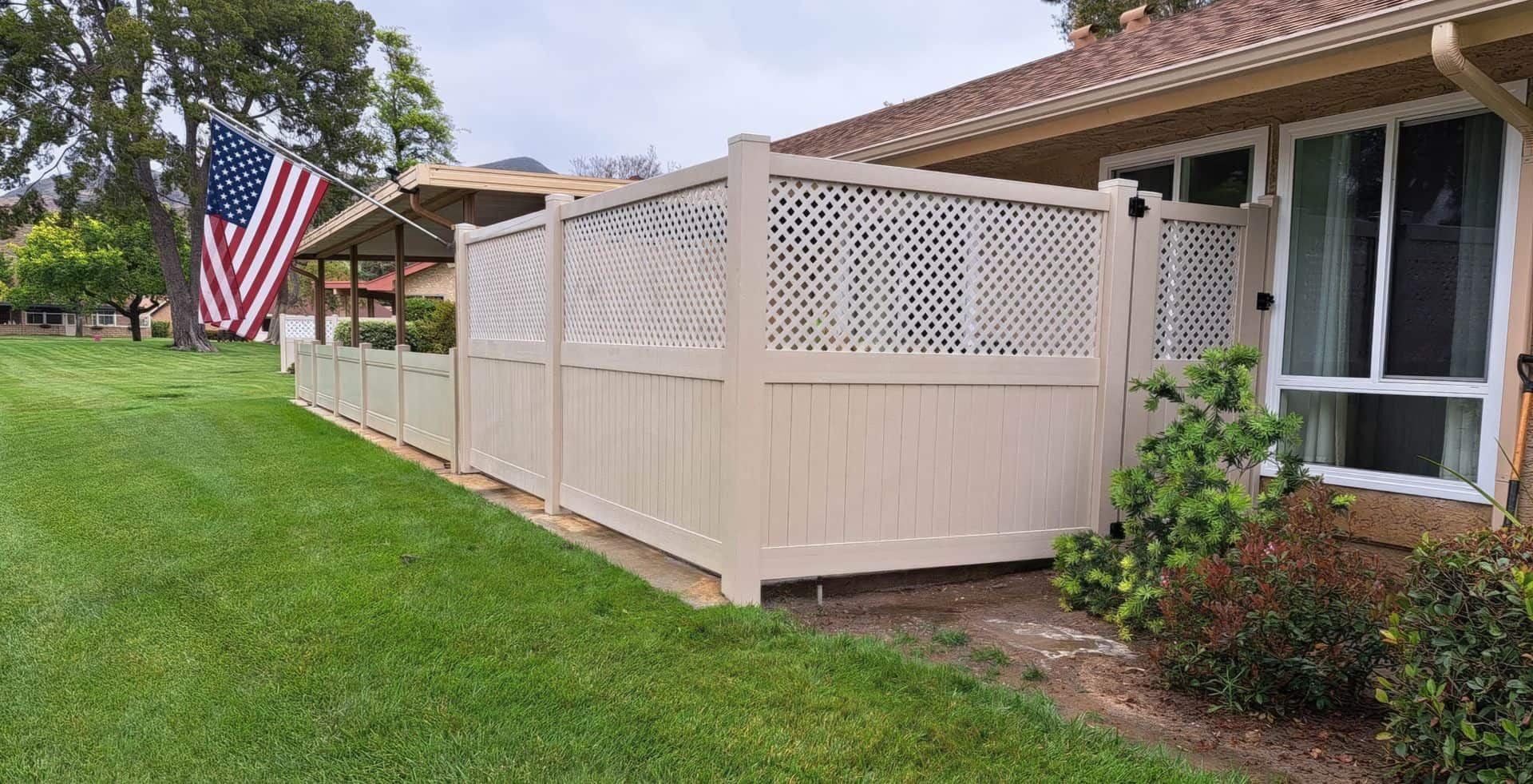 Tan vinyl fence beside small garden with bushes and shrubs and in front of grassy lawn.