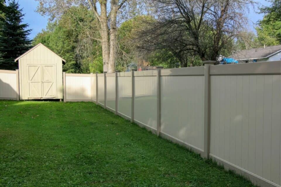 Vinyl clay colored fence right next to backyard shack in front of grassy lawn with trees in the back