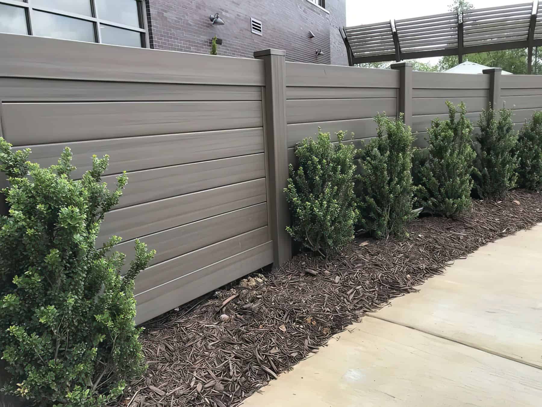 Vinyl clay colored fence surrounded leading to side passage wtih small shrubs lining concrete sidewalk in front.