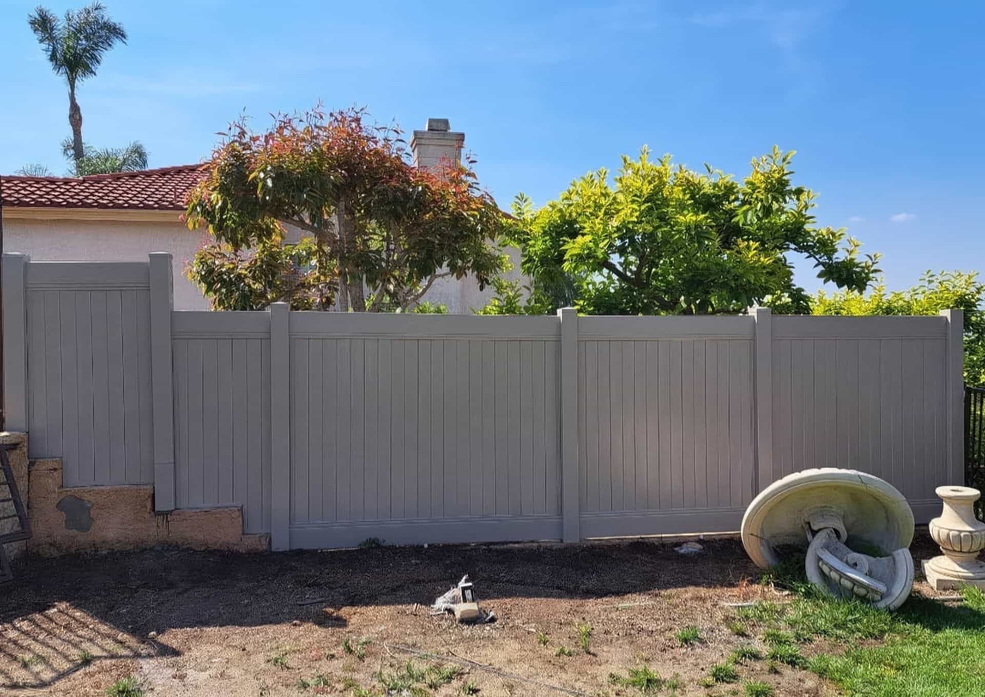 Vinyl clay colored fence with trees in the back and patchy green lawn in front with broken decorations