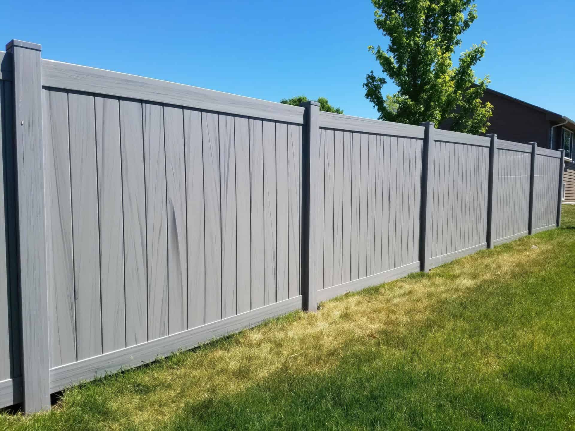 Vinyl coastal cedar colored fence in front of a grassy lawn and the with a large tree and blue sky in the background