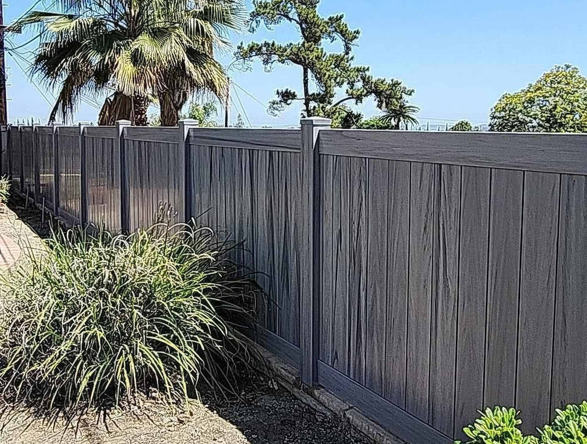 Vinyl coastal cedar colored fence in front of large palm tree and behind small bushes in backyard of suburban home