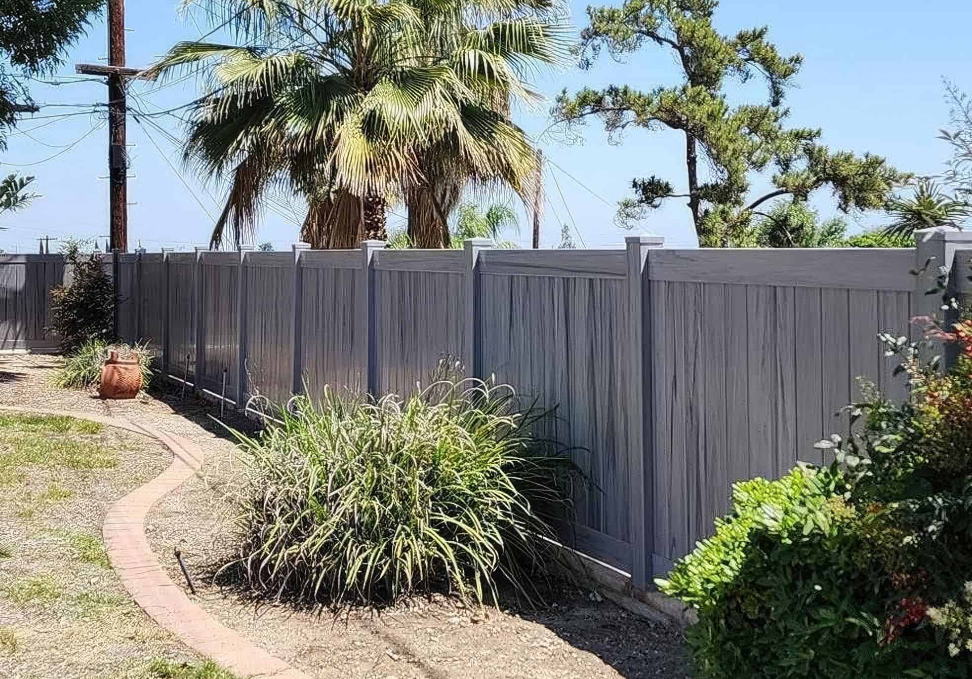 Vinyl coastal cedar colored fence creating boundary around the h backyard with dry grass lawn, small bushes and hedges, and trees in the background.