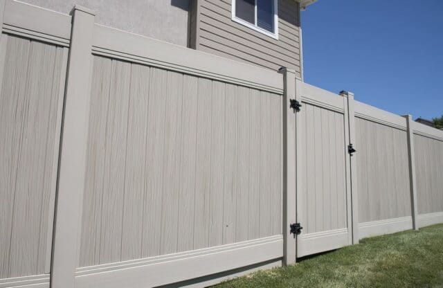 Vinyl driftwood colored fence with gate on grassy lawn on side entrance for beige colored suburban home.