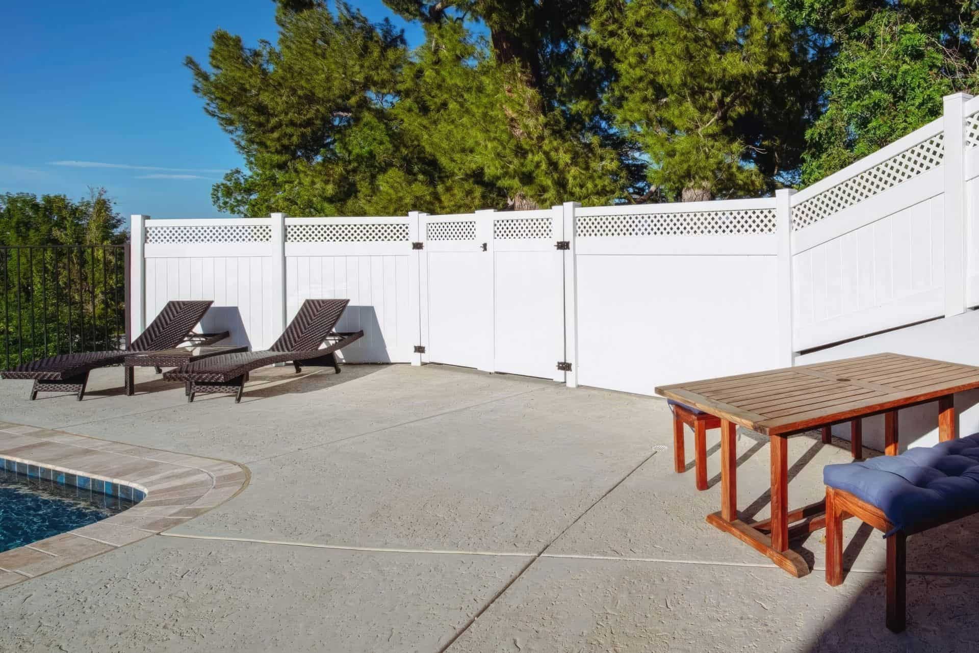 White vinyl fence surrounding modern suburban home in background with lounge chairs by the poolside.
