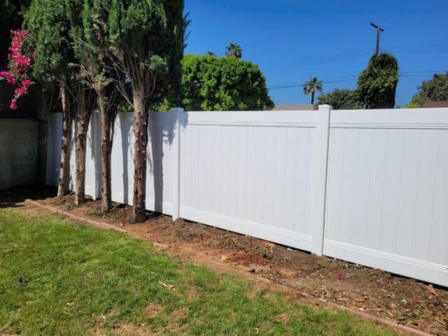 White vinyl fence surrounded by lush greenery, small plants, and trees in the backdrop, creating a serene and picturesque landscape.