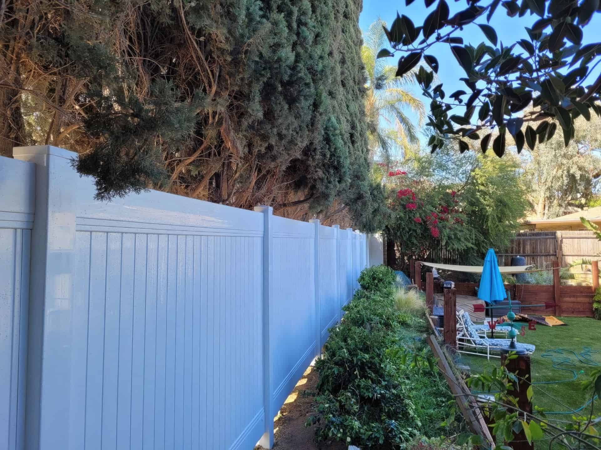 Large white vinyl fence bordering front lawn with trees and small bushes and separating it from concrete sidewalk