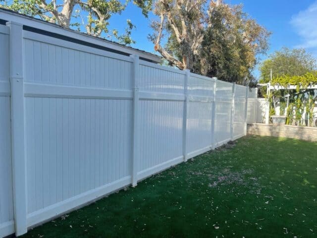 Large white vinyl fence separating two houses from the backyard with a small vertical garden and lush grass.