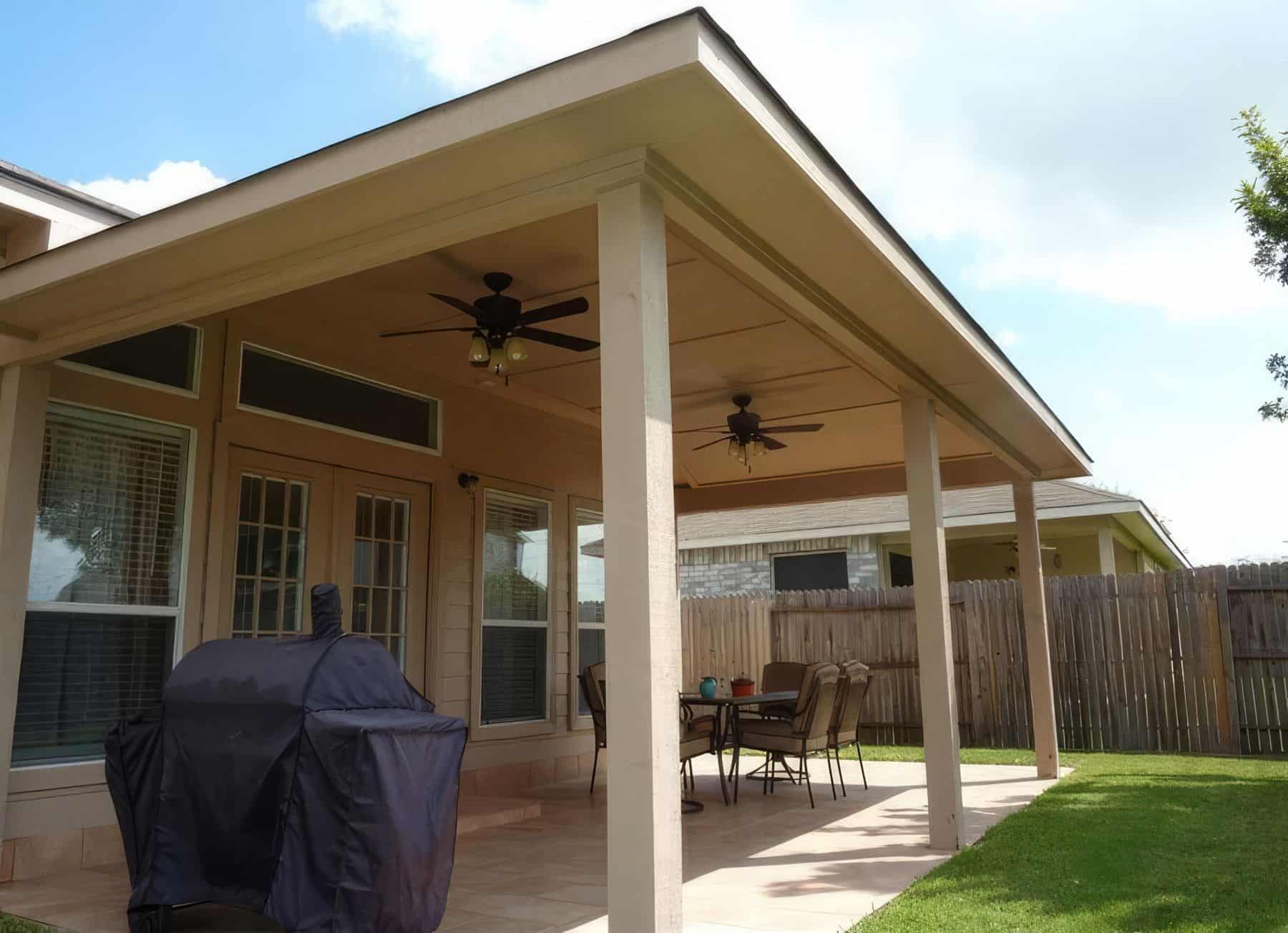 Outdoor furniture vinyl patio cover with ceiling fan, outdoor table & chairs on grassy lawn, surrounded by trees in the background.
