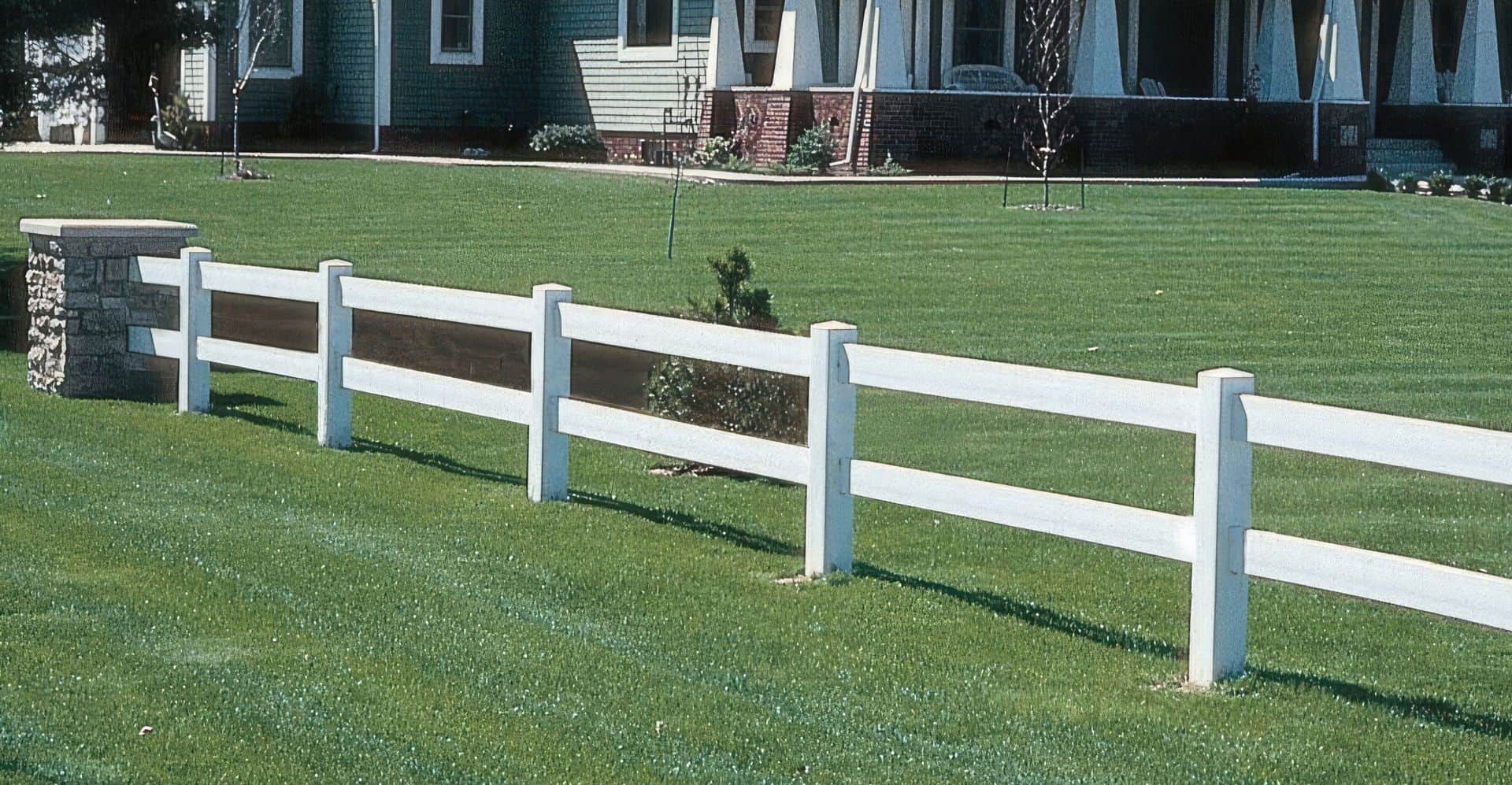 Vinyl 2-rail ranch fence in open field, urban setting. Suburban home with grassy lawn, trees in background.