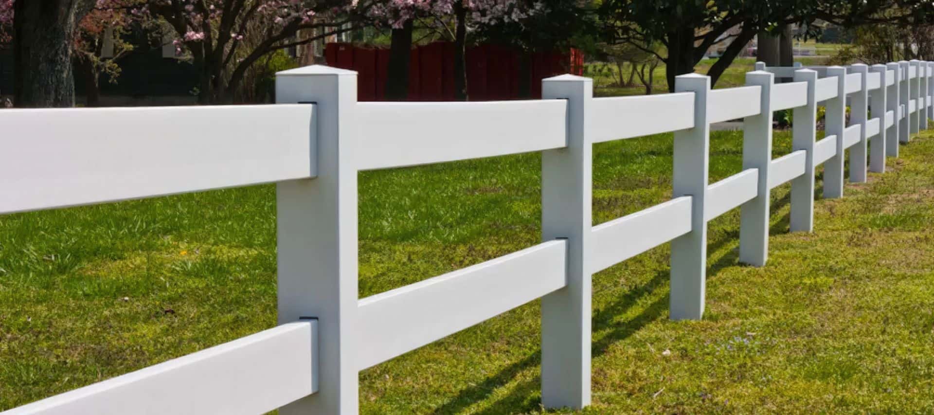 Vinyl 2-rail ranch fence in urban setting. Open field with grassy lawn, trees in the background. Serene and secure landscape.