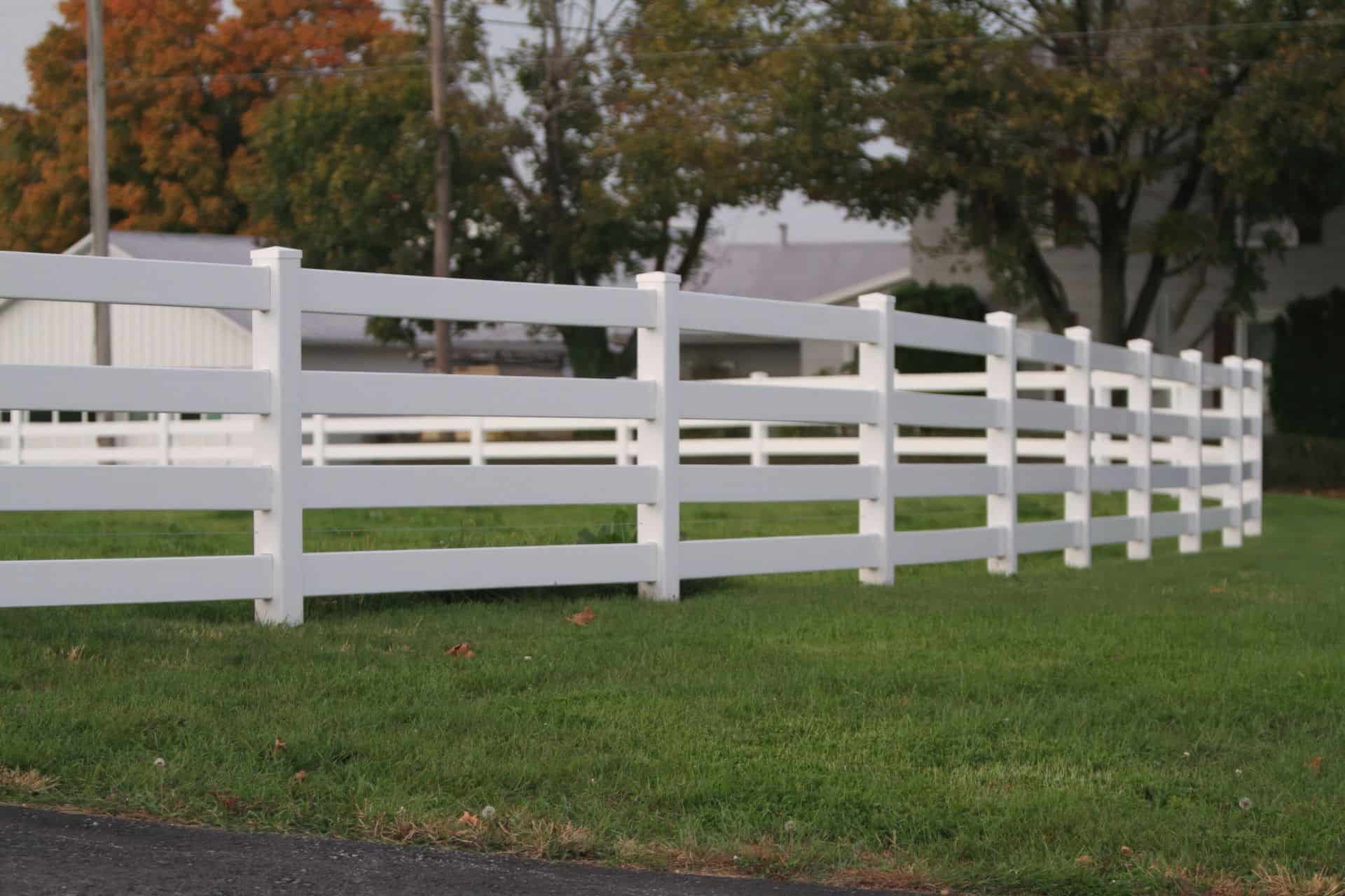 Country home with vinyl 4-rail ranch fence, front lawn, open field, and trees in the background.