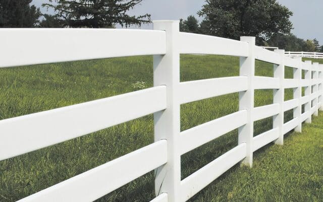 Vinyl 4-rail ranch fence under clear sky, with lush grassy lawn & trees in the background. Idyllic outdoor setting.