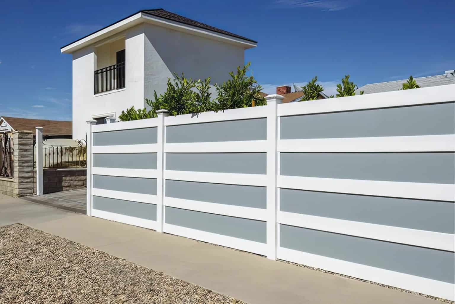 Vinyl & acrylic privacy fence beside concrete sidewalk, under clear sky, with trees in the background.