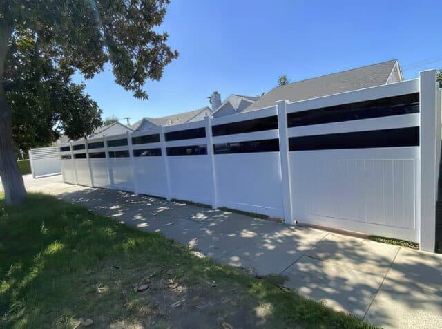 Vinyl & acrylic privacy fence encloses grassy lawn & concrete sidewalk with houses in background. Scenic suburban setting.