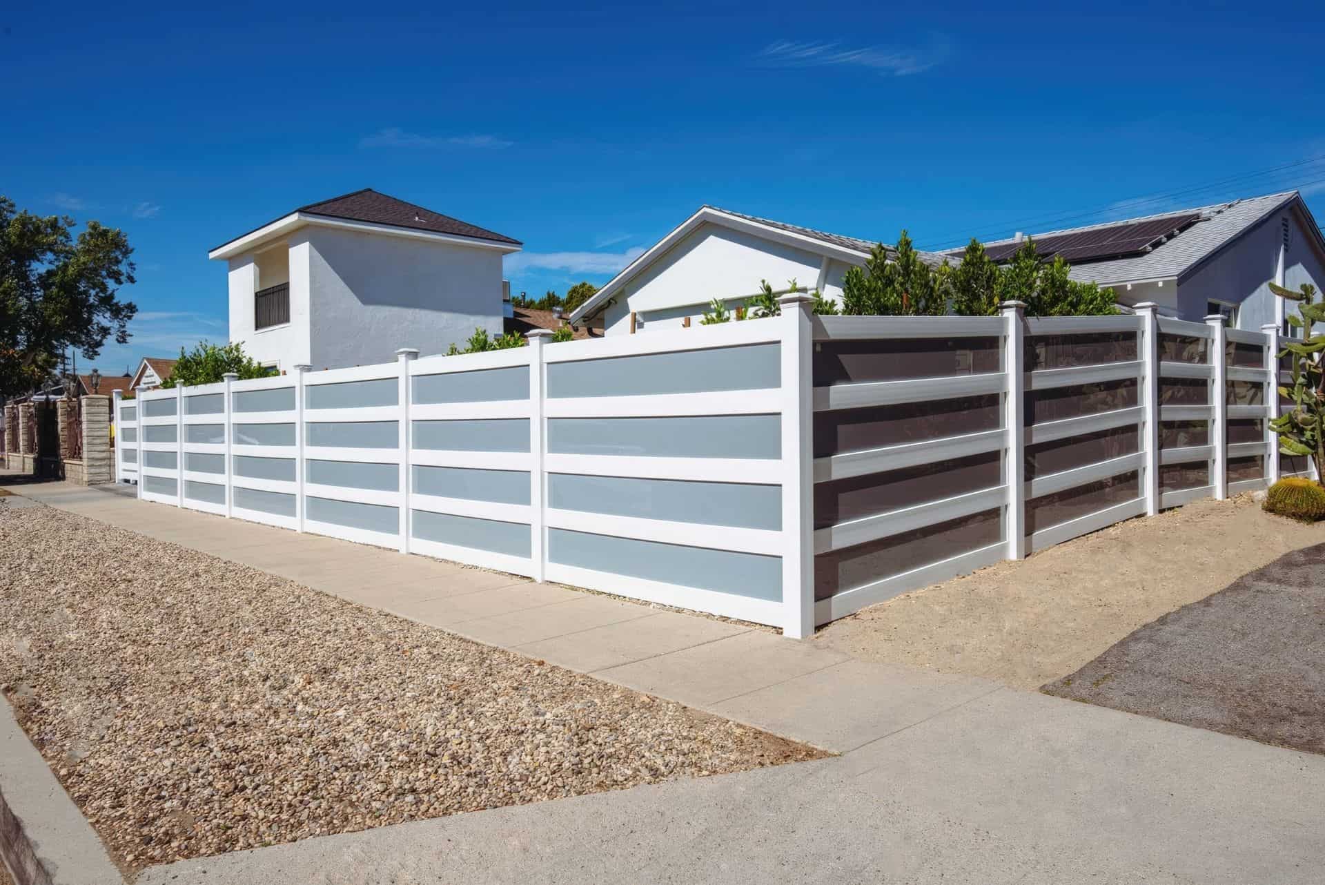 Vinyl & acrylic privacy fence by a concrete sidewalk, offering increased privacy with trees and houses in the background.