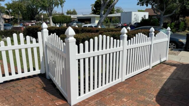 Vinyl arched picket fence & fence gate alongside a brick driveway. Trees in the background and concrete sidewalk.