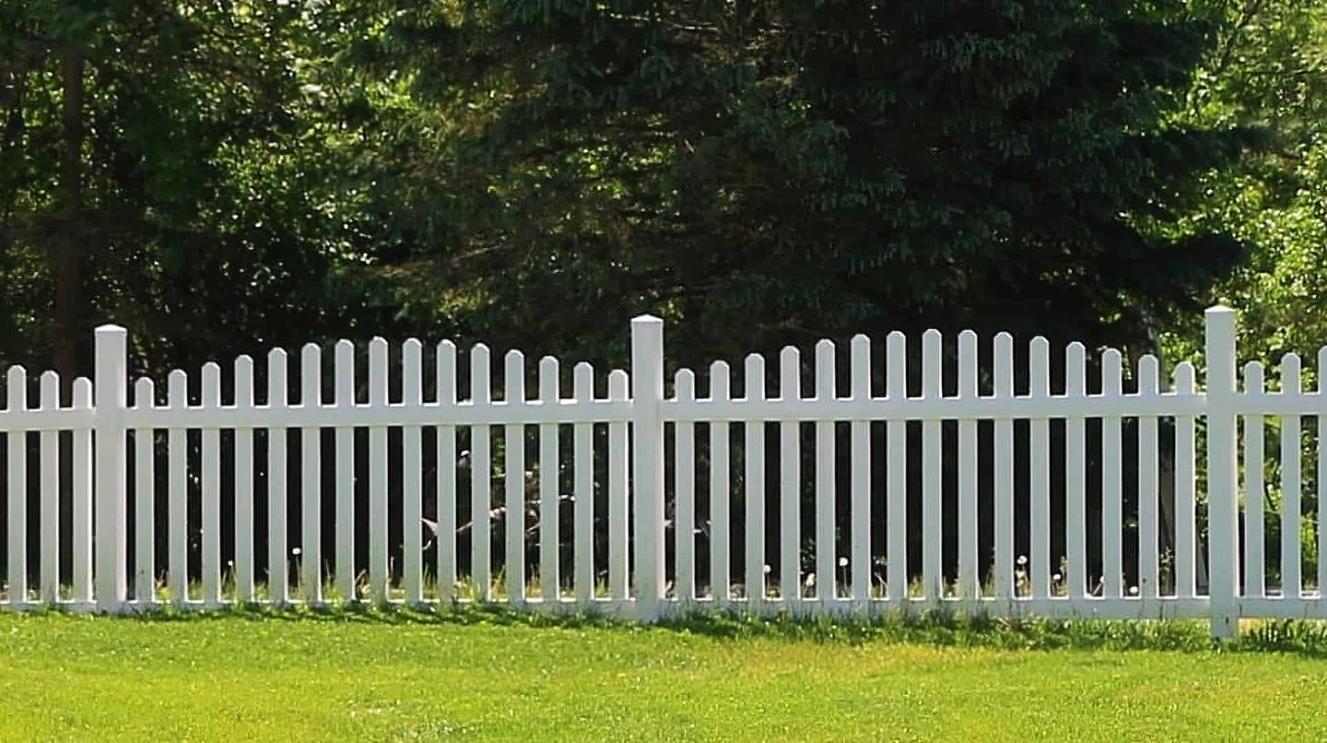 Vinyl arched picket fence bordering a grassy lawn with a small garden and trees in the background.