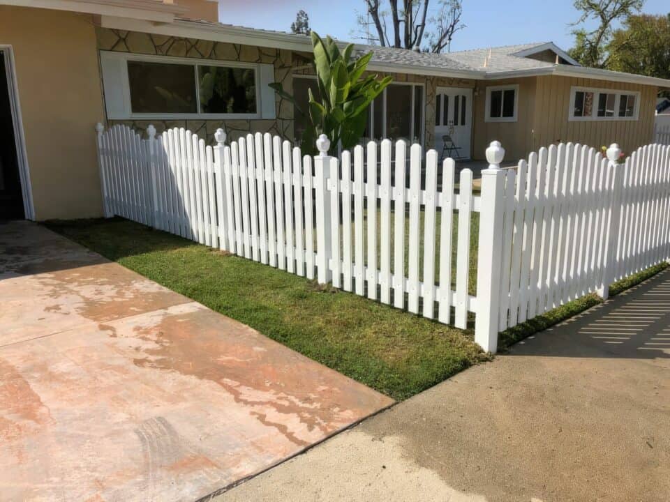 Charming vinyl arched picket fence surrounding small garden, with trees in the background, alongside a concrete driveway.