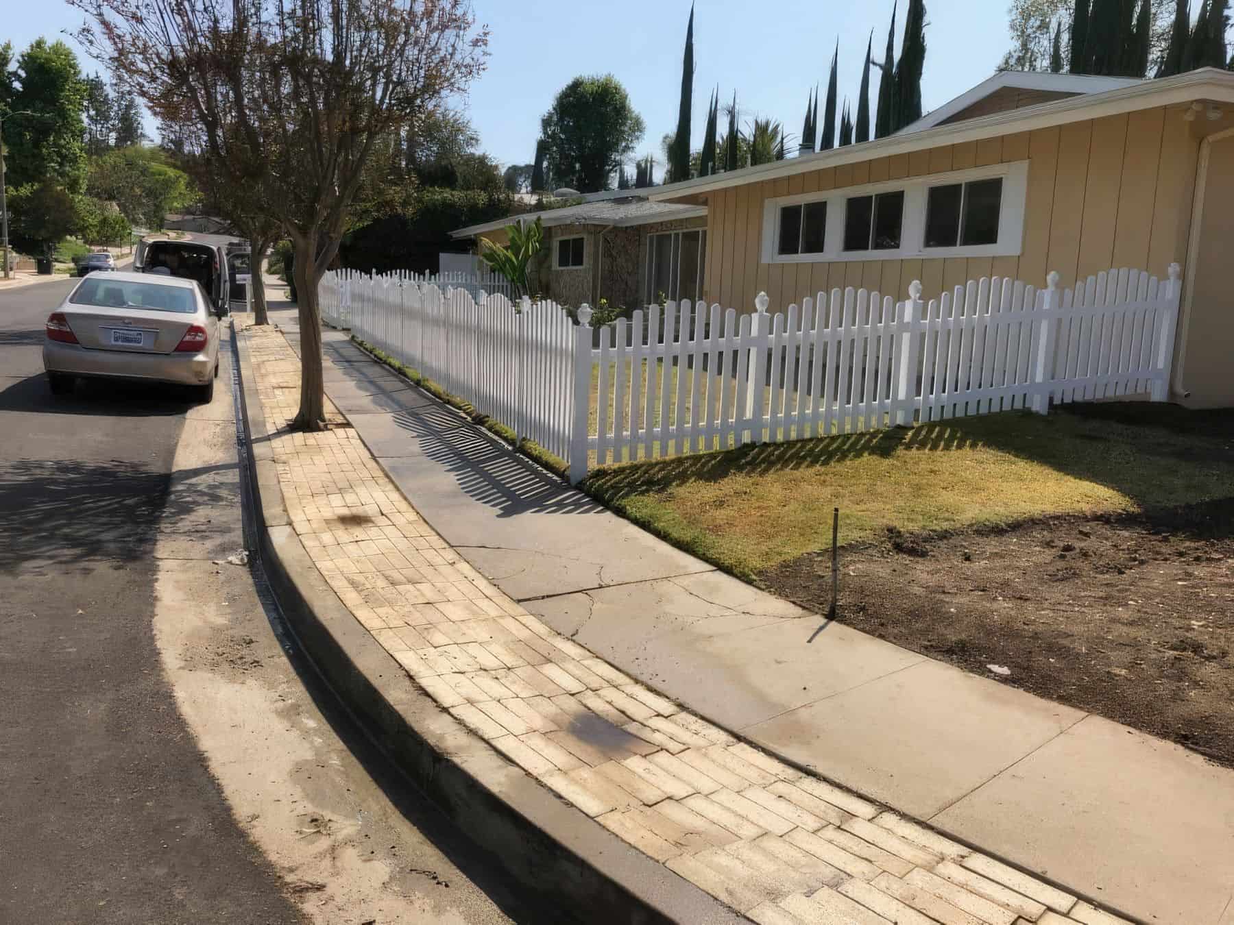 Vinyl arched picket fence surrounds small garden, trees in background & concrete sidewalk add charm to the scene.