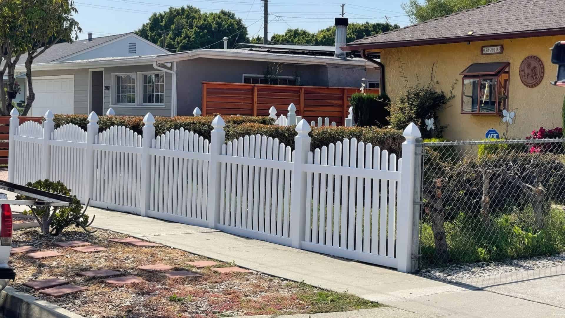 Vinyl arched picket fence & brick driveway beside a small garden & concrete sidewalk. Charming houses in the background.