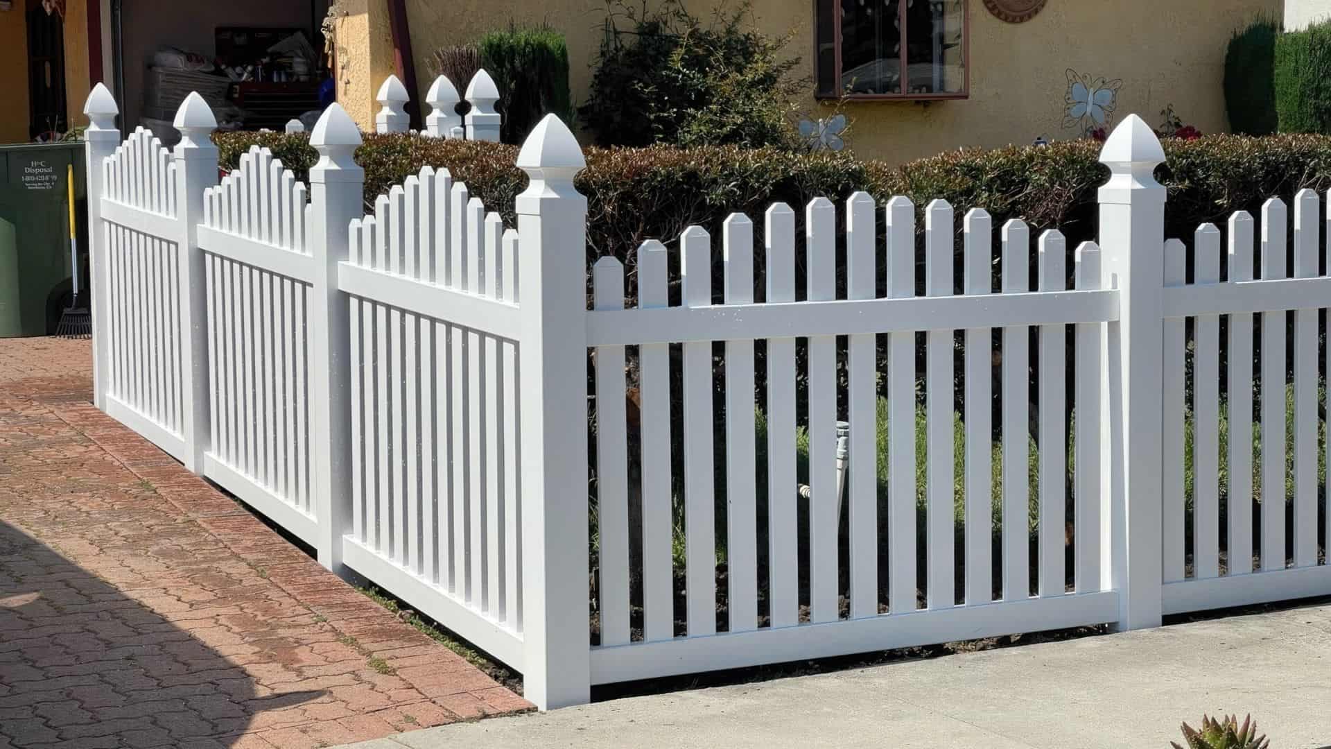 Vinyl arched picket fence beside concrete sidewalk, brick driveway & charming houses in the background.