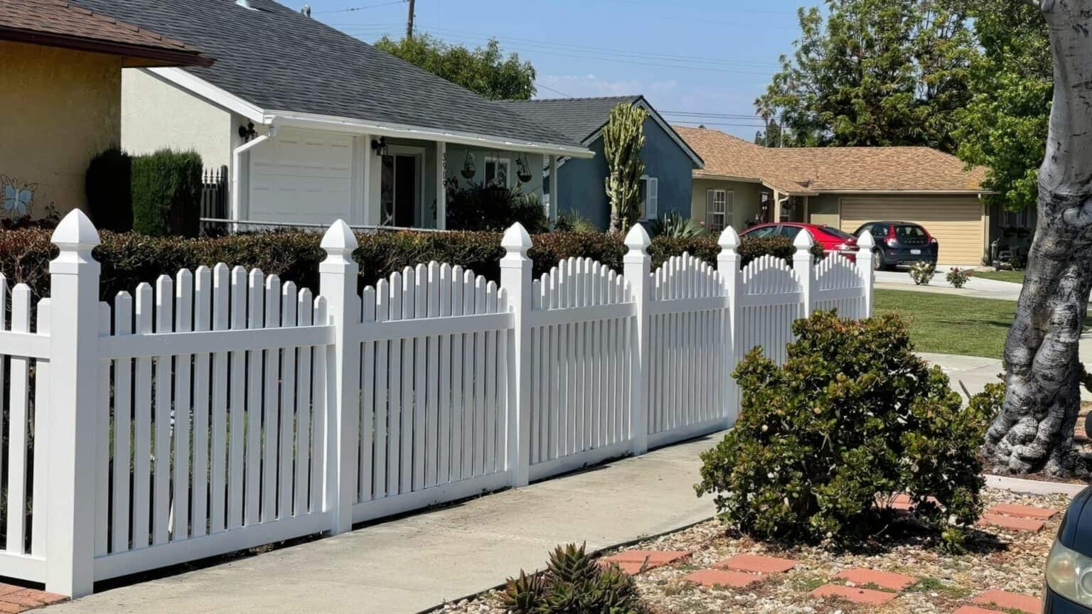 Vinyl arched picket fence & brick driveway. Houses stand in the background as a concrete sidewalk completes the scene.