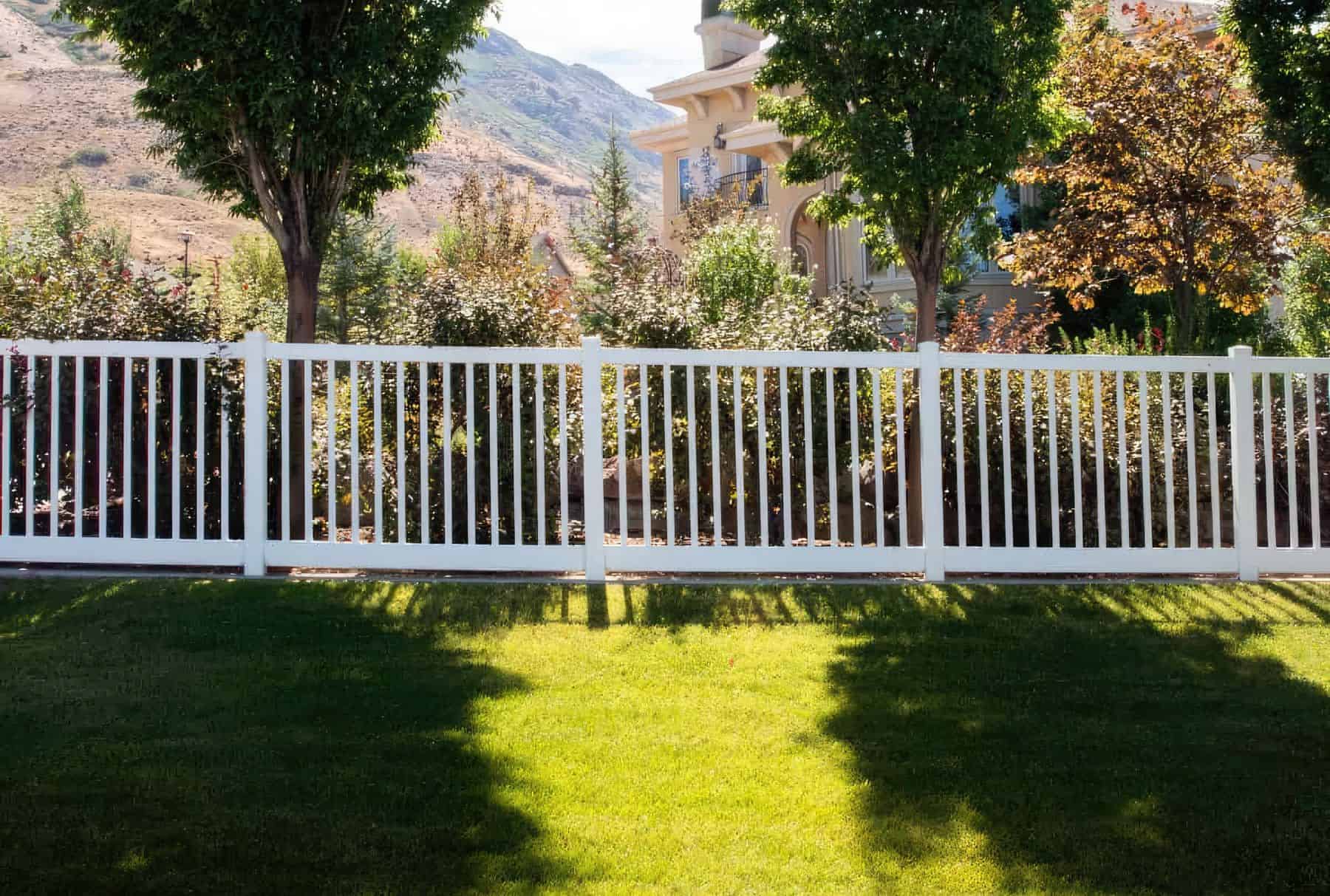 Vinyl closed-top picket fence surrounding garden on grassy lawn with trees in background - a charming outdoor oasis.