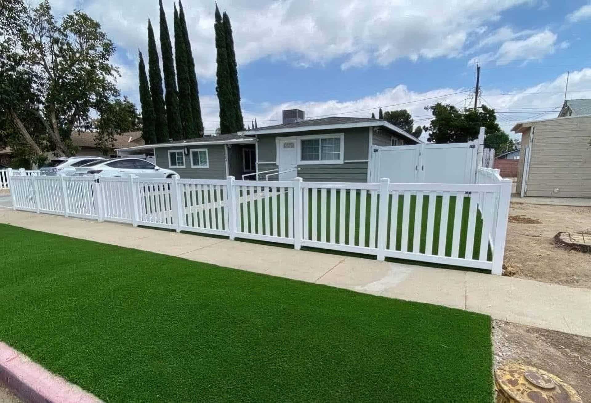 Suburban house surrounded by vinyl closed-top picket fence, lush lawn & trees, with a concrete sidewalk in the foreground.