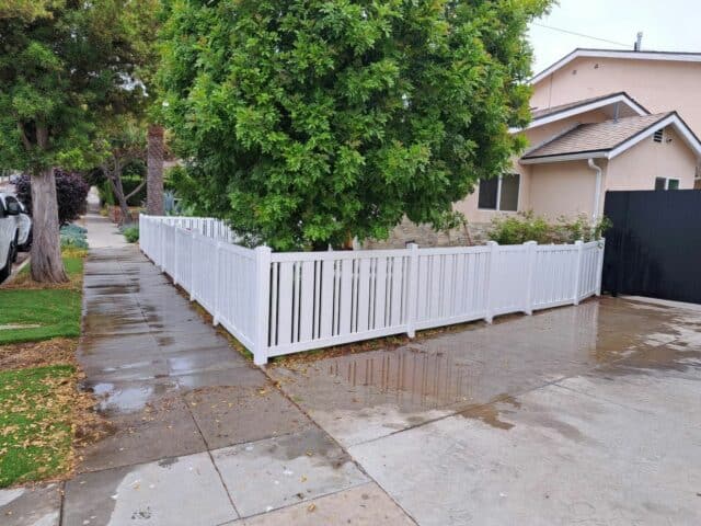 Vinyl closed-top picket fence with front lawn, concrete driveway, sidewalk, and trees in the background.