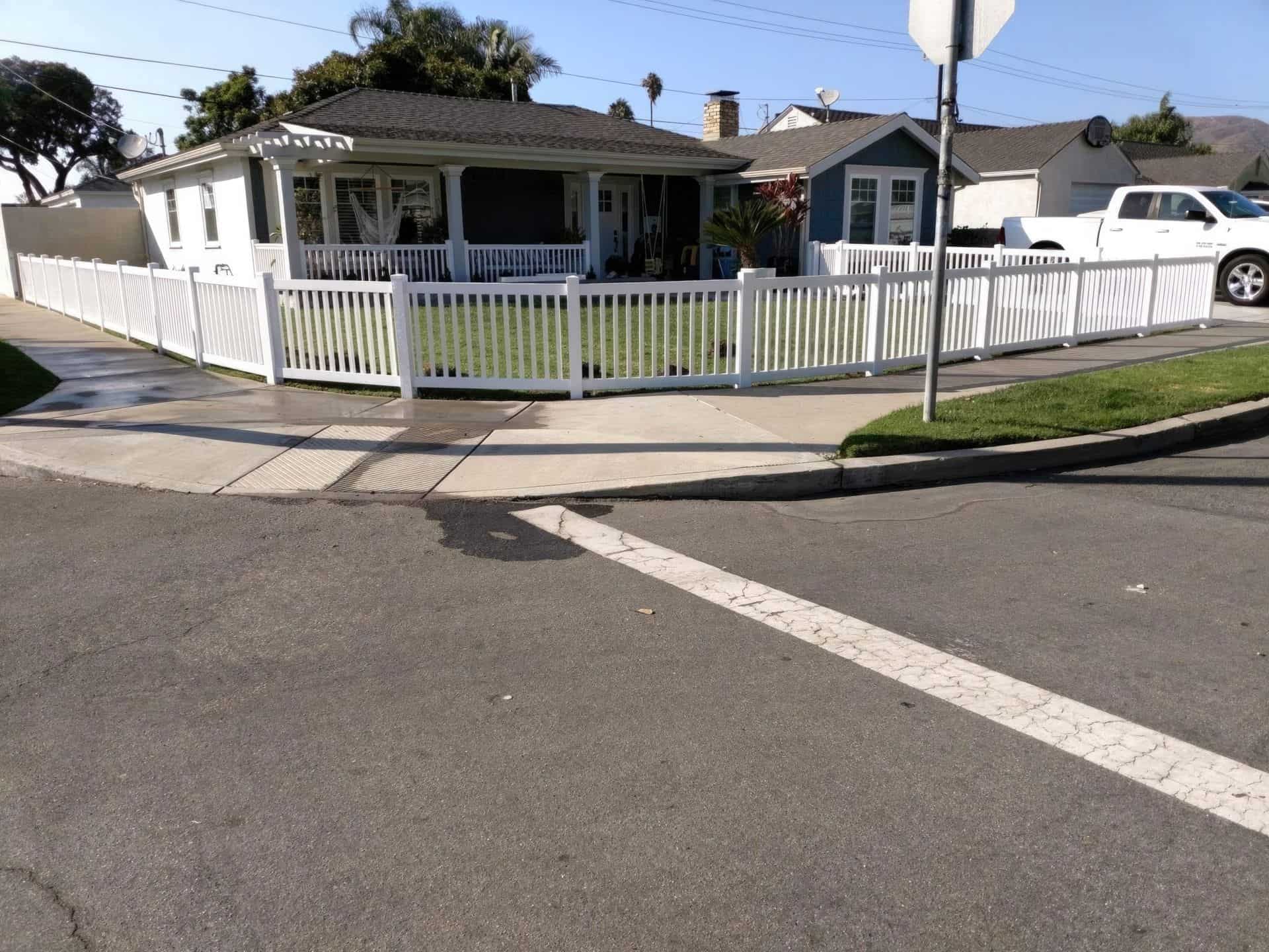 Suburban house with vinyl closed-top picket fence, concrete driveway, sidewalk, and trees in the background.