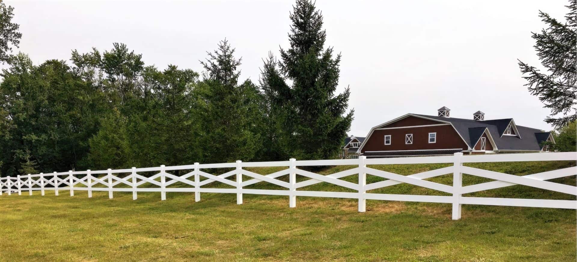 Vinyl cross rail ranch style fence creating boundary around open field with trees in the background as well as farmhouses.