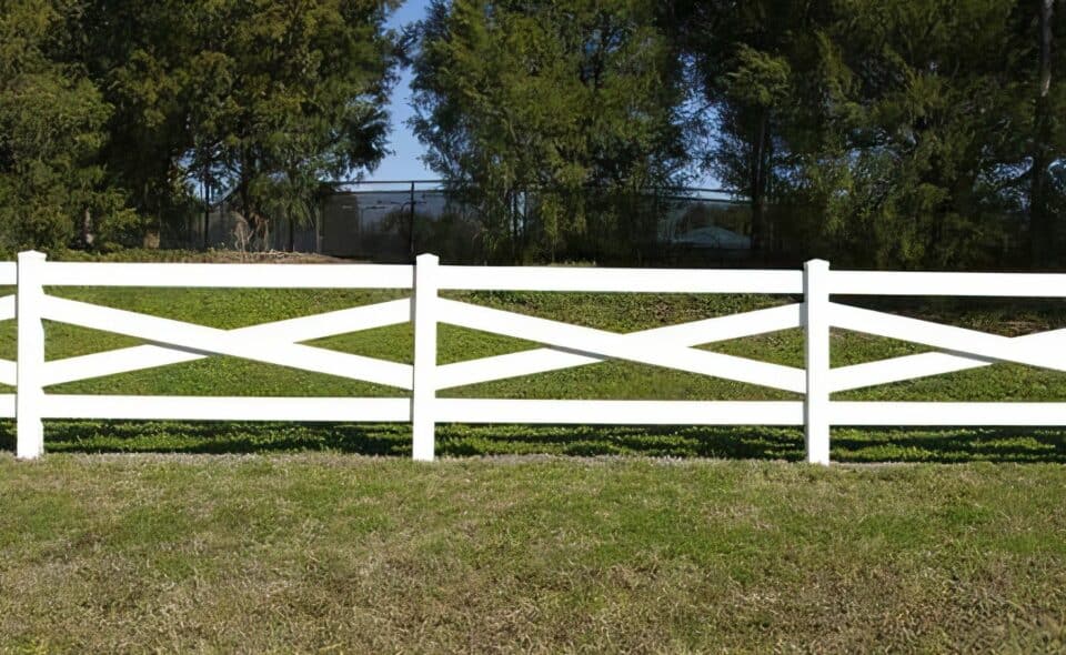 Vinyl cross rail ranch style fence with trees & grassy lawn in the background, creating a serene outdoor setting.