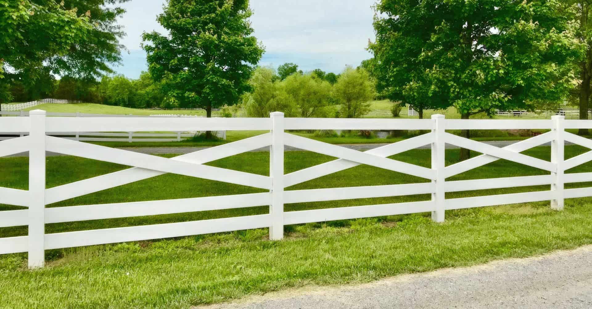 Vinyl cross rail ranch style fence surrounded by lush grass and tall trees in the background - idyllic countryside scene.