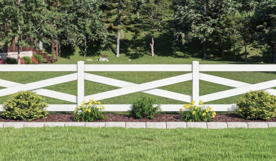Vinyl cross rail ranch style fence surrounded by lush lawn, small plants, and distant trees, creating a serene outdoor setting.