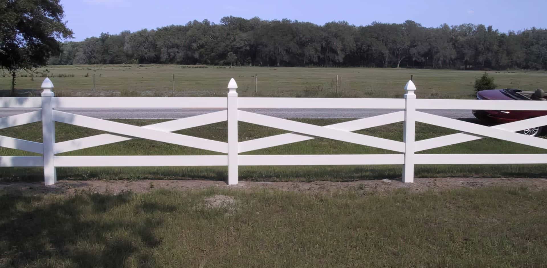 Vinyl cross rail ranch style fence on private grassy lawn far away from road with many trees in the background