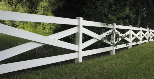 Vinyl cross rail ranch style fence on grassy lawn surrounded by trees and winding roads in the distance.