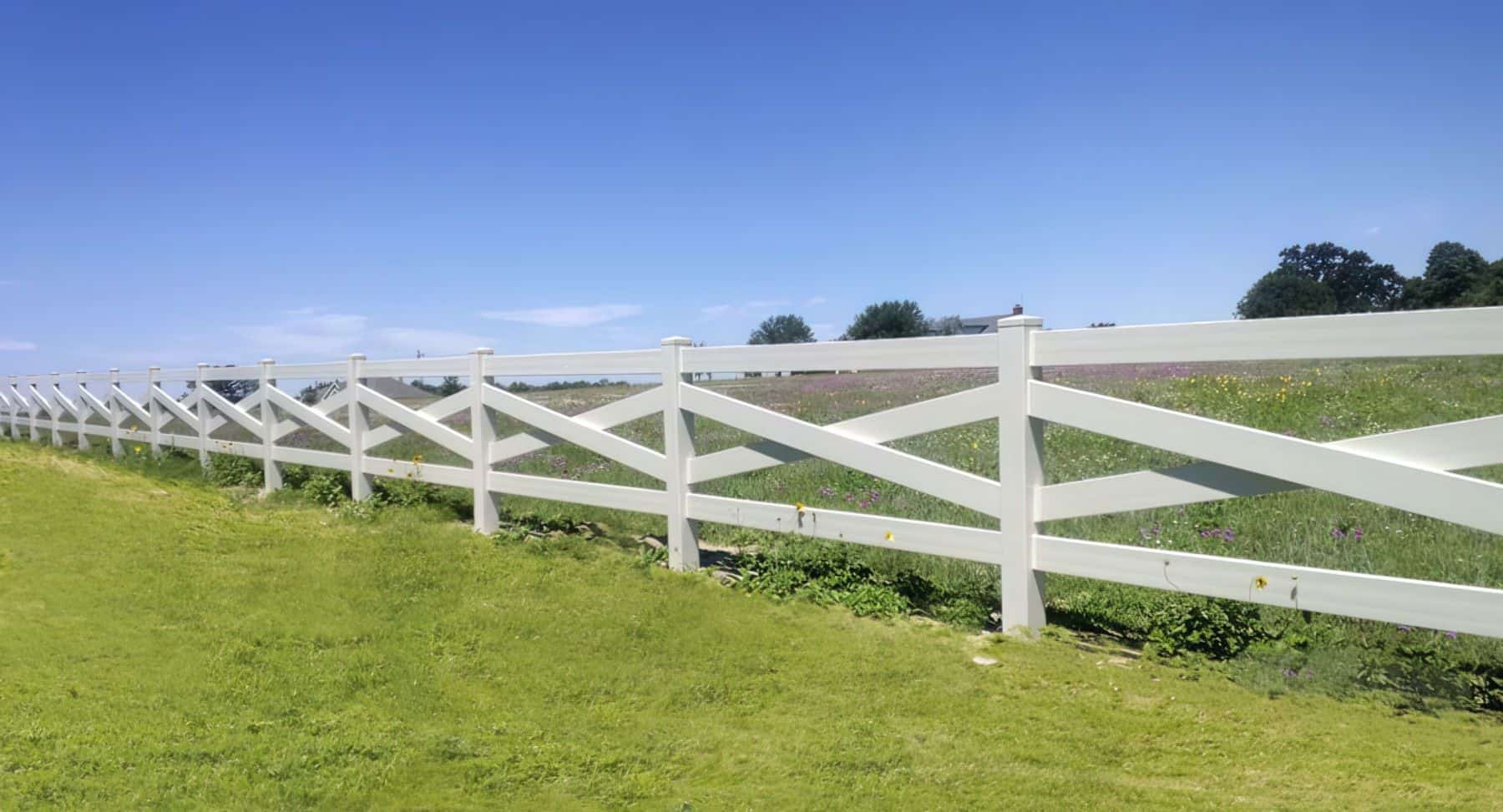 Vinyl cross rail ranch style fence surrounding grassy lawn, with trees in the background - picturesque countryside scene.