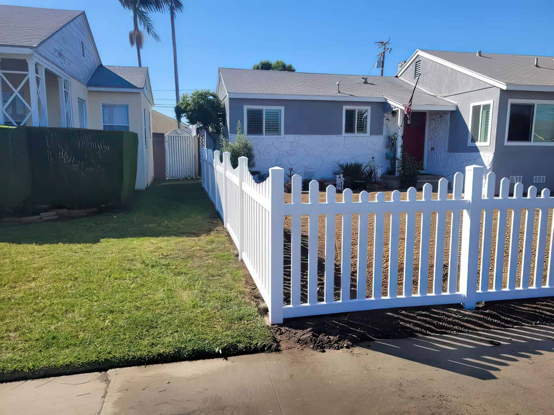 Vinyl dog ear picket fence surrounds ranch house with concrete sidewalk & driveway; suburban homes in background.
