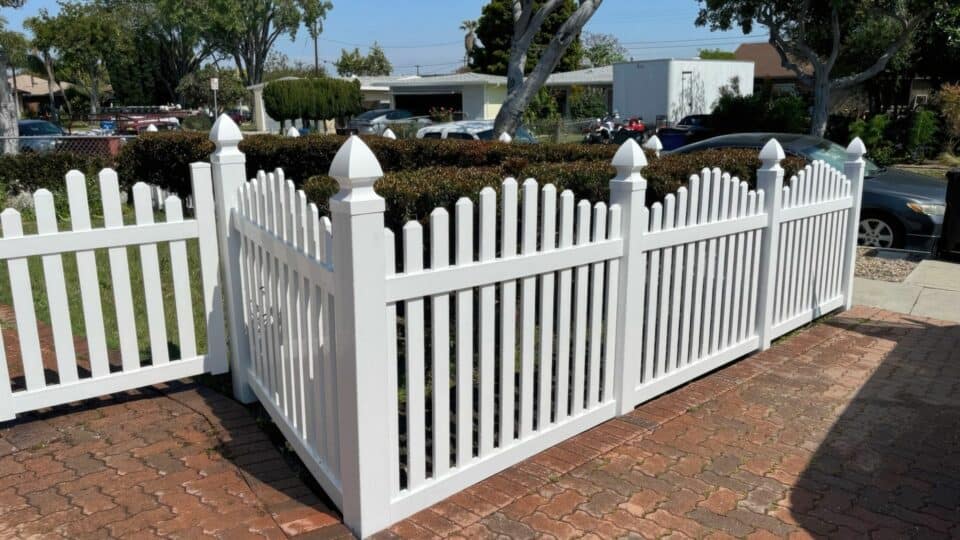 Vinyl dog ear picket fence surrounding suburban house with concrete sidewalk & green front lawn.