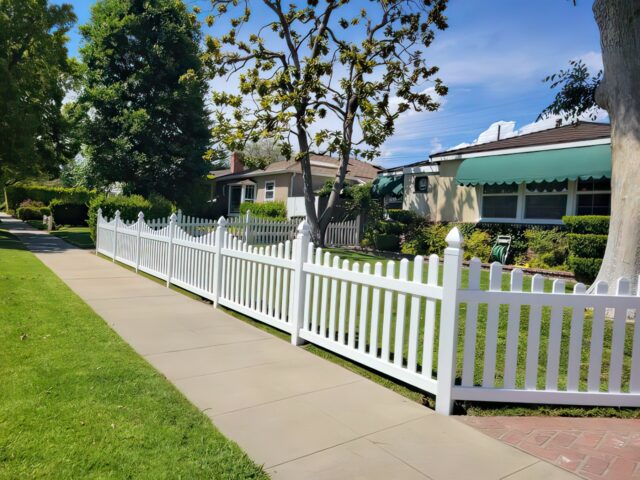 A vinyl dog ear picket fence surrounding front lawn with bigger trees in the background and small hedges in the garden